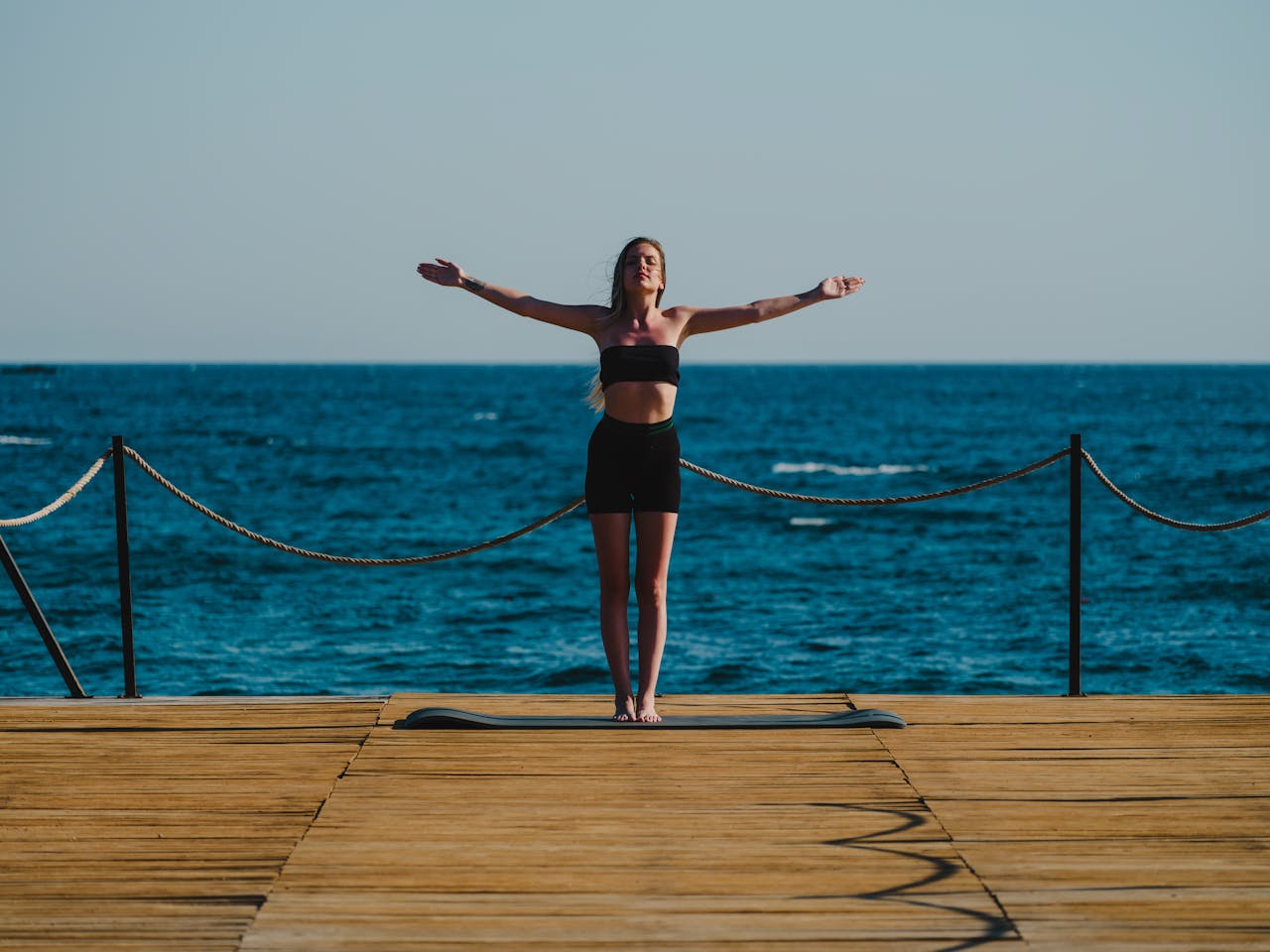 Home A woman in activewear stands in a yoga pose on a wooden dock near the sea, embracing a tranquil moment.